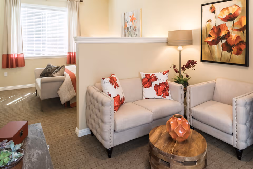 Cozy living area with beige loveseat and armchairs, floral pillows, a wooden coffee table and artwork, separated from a bed by a half wall.