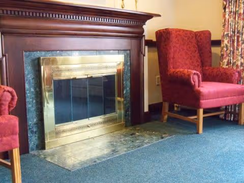 A cozy interior corner featuring a wooden fireplace with a brass-framed glass door, flanked by two red upholstered armchairs on a blue carpeted floor, with floral curtains in the background.