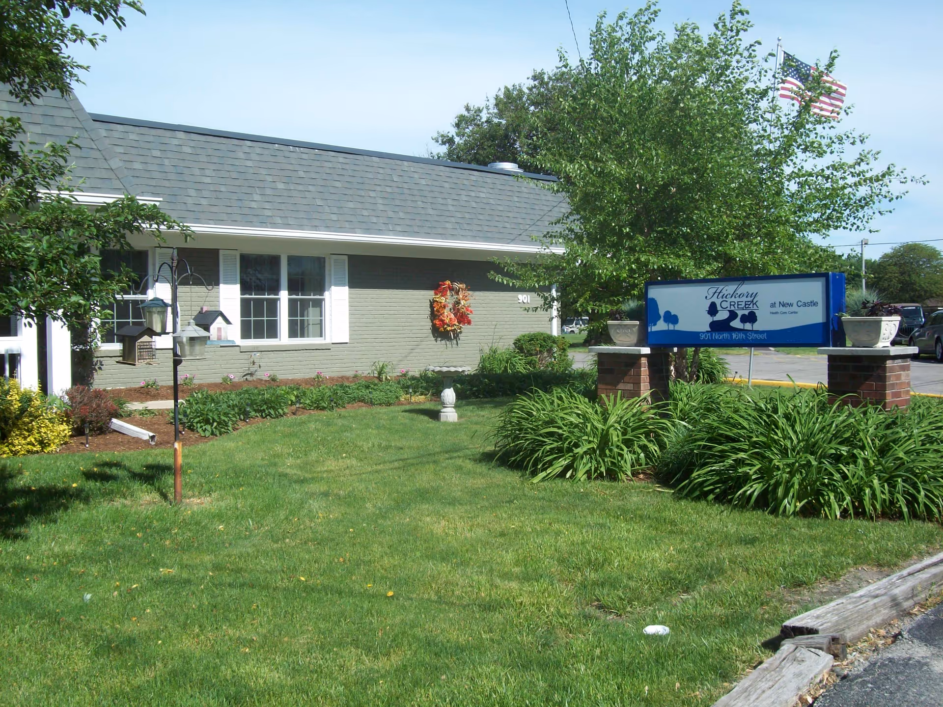 Exterior view of a single-story building with a gray shingled roof and light green brick walls. There is a well-maintained lawn with shrubs and plants in front of the building. A sign reading 'Hickory Creek at New Castle' is displayed on brick pillars surrounded by greenery. An American flag is flying on a flagpole behind the sign.