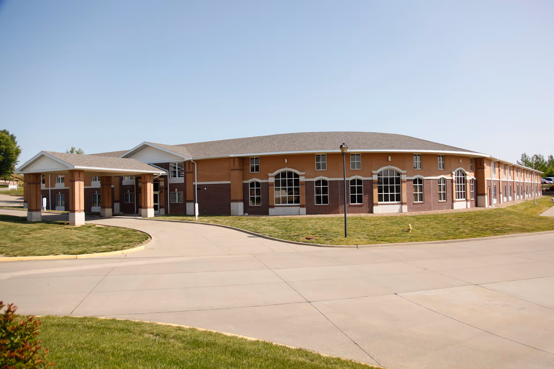 Exterior view of a large two-story brick building with multiple windows and a covered entrance, surrounded by a paved driveway and green lawn under a clear blue sky.