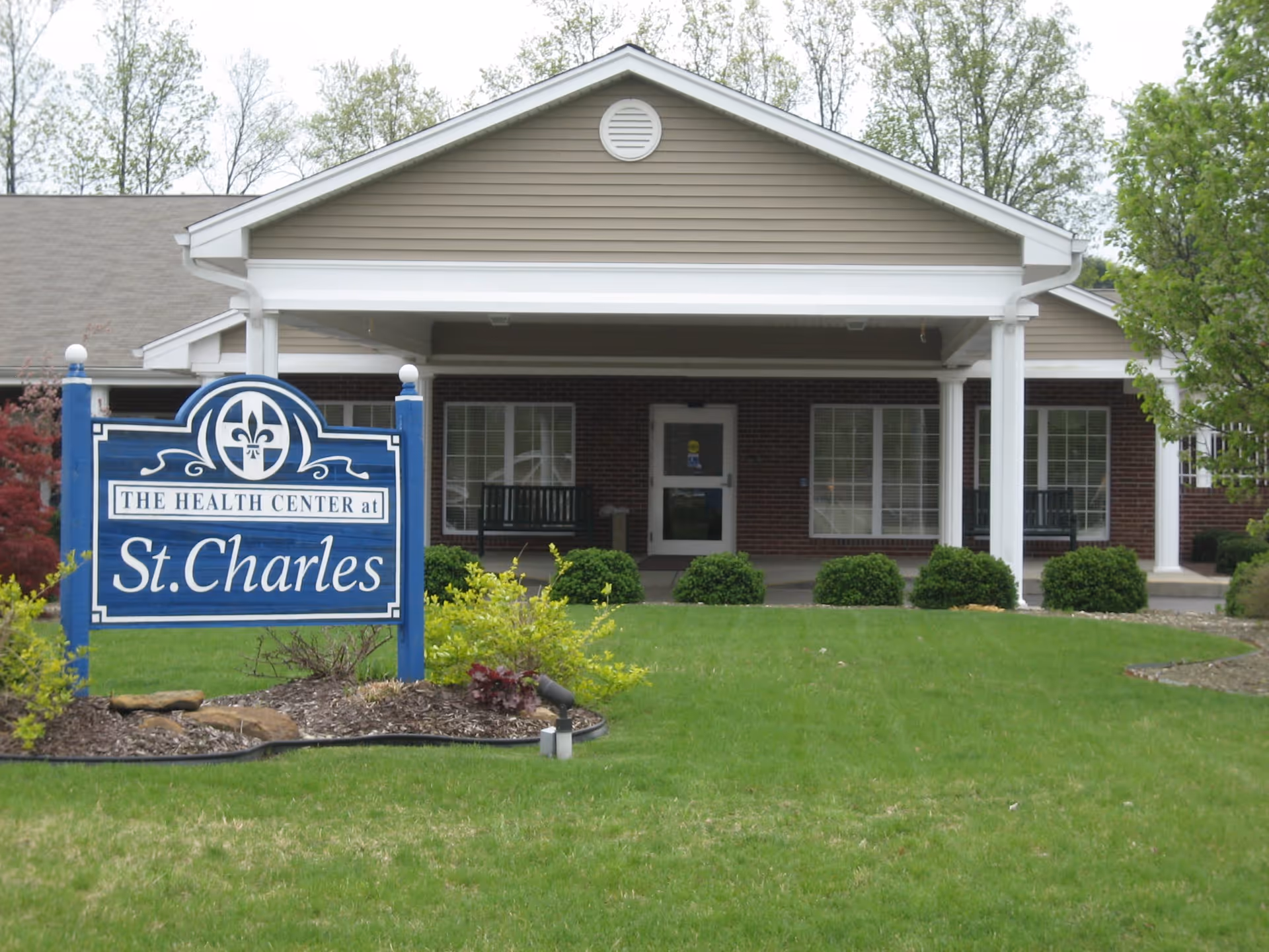 Exterior view of The Health Center at St. Charles, showing a single-story building with a covered entrance, large windows, and a well-maintained lawn with shrubs and small trees. A blue sign with white text is prominently displayed in front of the building.