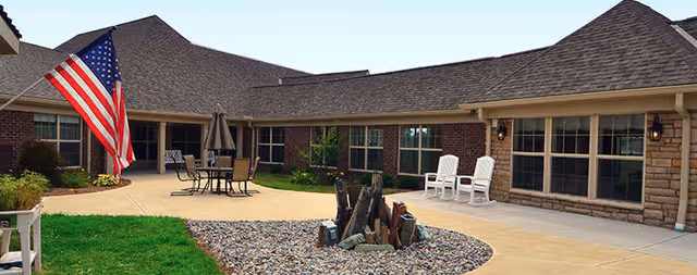 Outdoor courtyard area of a senior living facility with a paved walkway, a small rock garden with decorative logs, an American flag on a pole, patio tables with umbrellas and chairs, and two white rocking chairs near the building's windows.