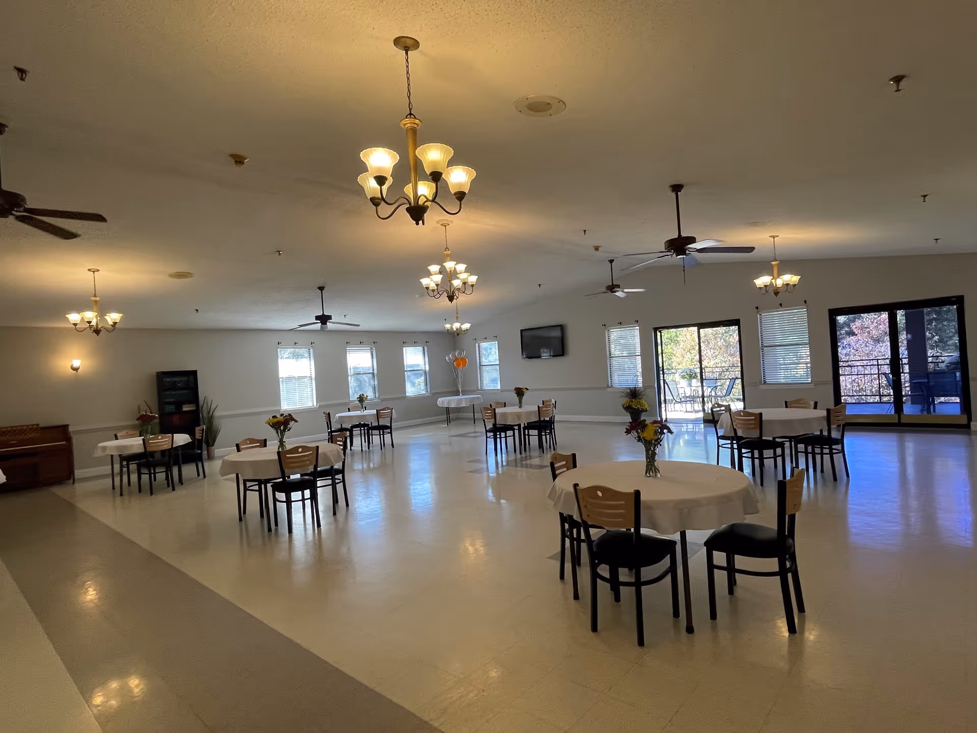 A spacious dining room with several round tables covered with white tablecloths, each surrounded by wooden chairs. The tables have small flower arrangements. The room has multiple ceiling fans and chandeliers providing warm lighting. Large windows and glass doors let in natural light and offer a view of an outdoor patio area with seating. The walls are light-colored, and there is a television mounted on one wall.