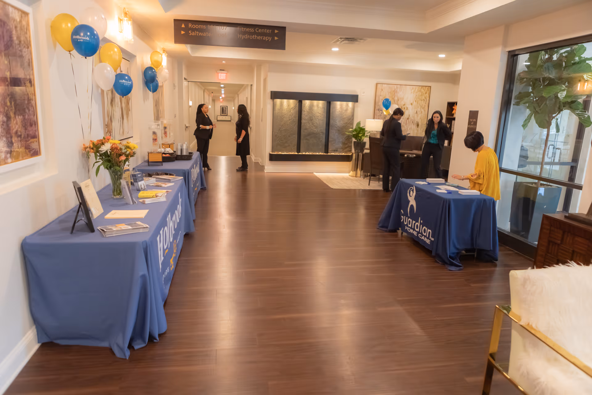 Interior hallway of a senior living facility with wooden flooring and tables covered with blue tablecloths displaying informational materials and balloons. Several people are standing and conversing near the tables. There is a water feature on the far wall and large windows on the right side letting in natural light.