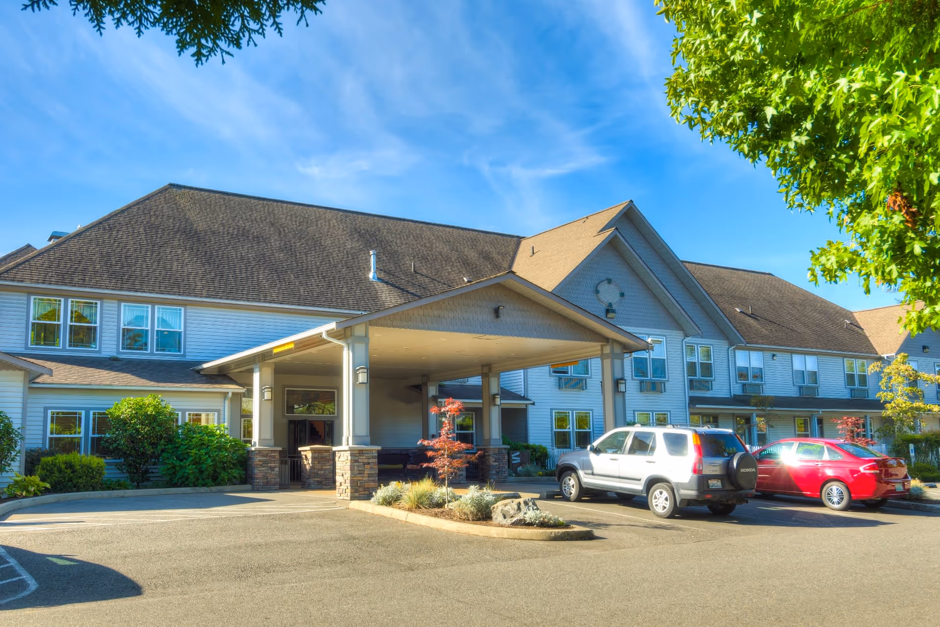 Exterior view of a senior living facility building with a covered entrance and several parked cars in front. The building has multiple windows, a sloped roof, and is surrounded by greenery including trees and bushes under a clear blue sky.