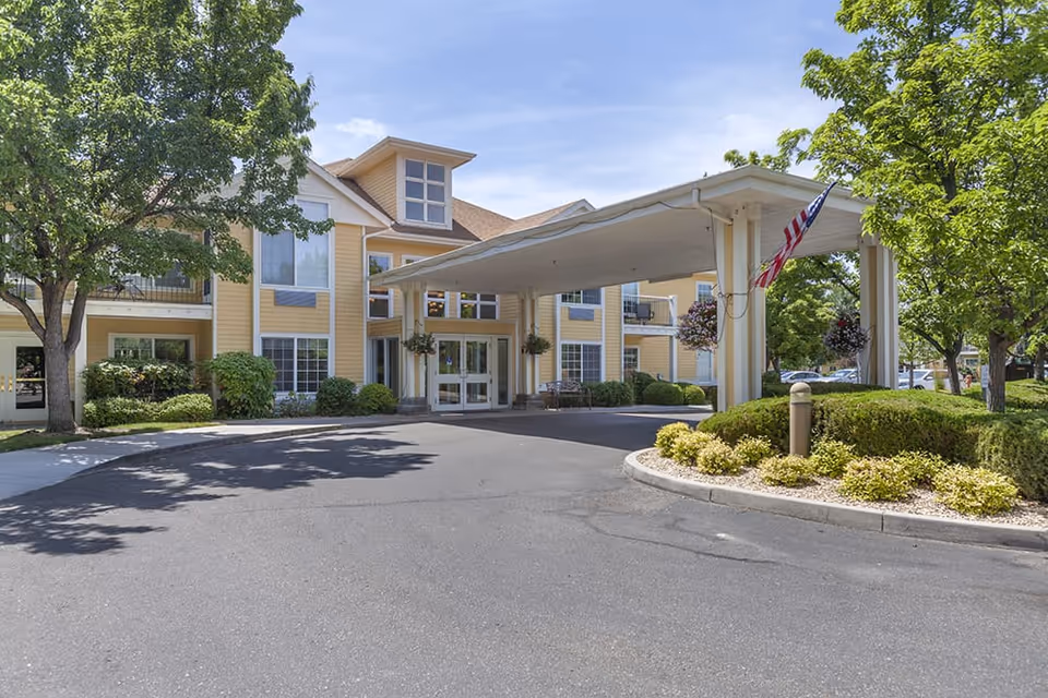 Exterior view of a senior living facility with a covered entrance, yellow siding, multiple windows, and surrounding greenery including trees and bushes under a clear blue sky.