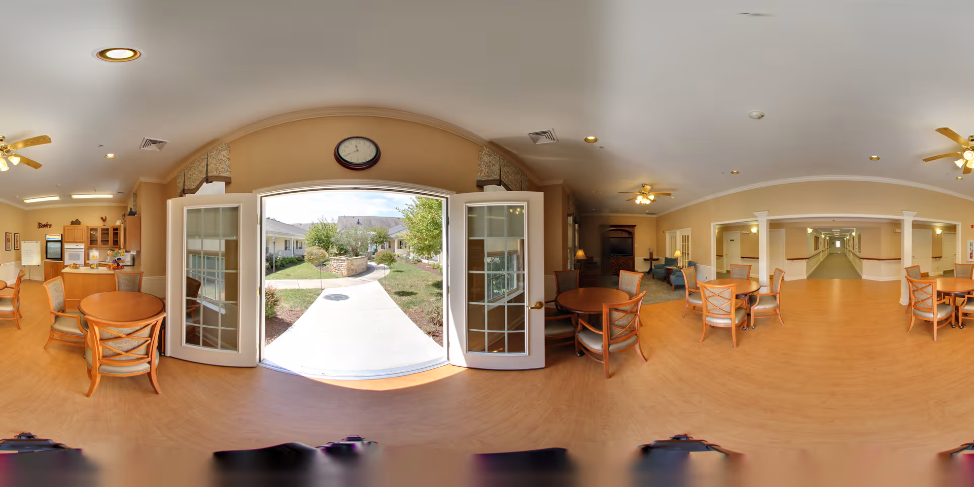 A spacious common area in a senior living facility with round wooden tables and chairs arranged on a light wood floor. Ceiling fans with lights are mounted on the ceiling. French doors open to a sunny outdoor garden area with a paved walkway and greenery. The room has beige walls and a clock above the doors. A hallway extends from the right side of the room.