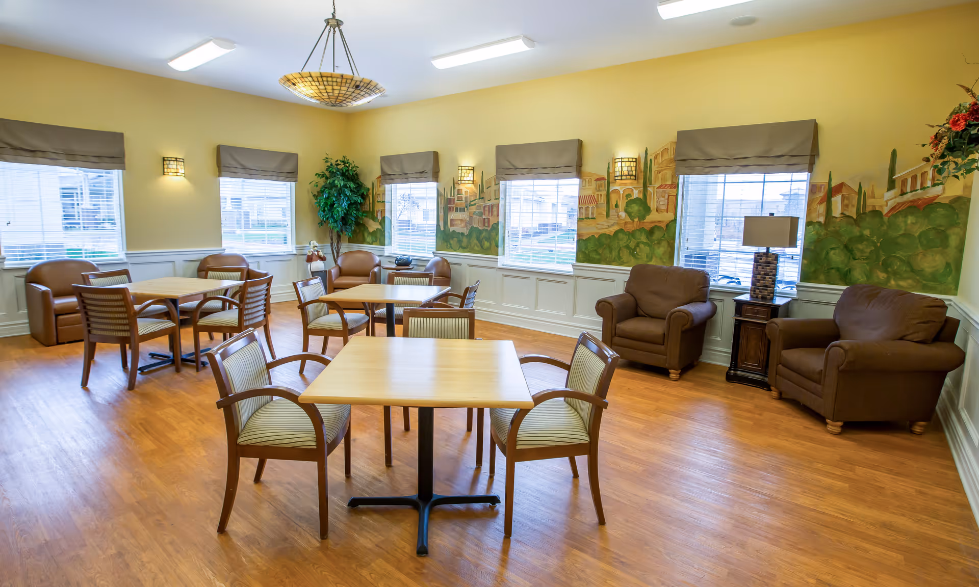 Bright communal room with several square dining tables and chairs, upholstered armchairs, windows, and a painted wall mural.
