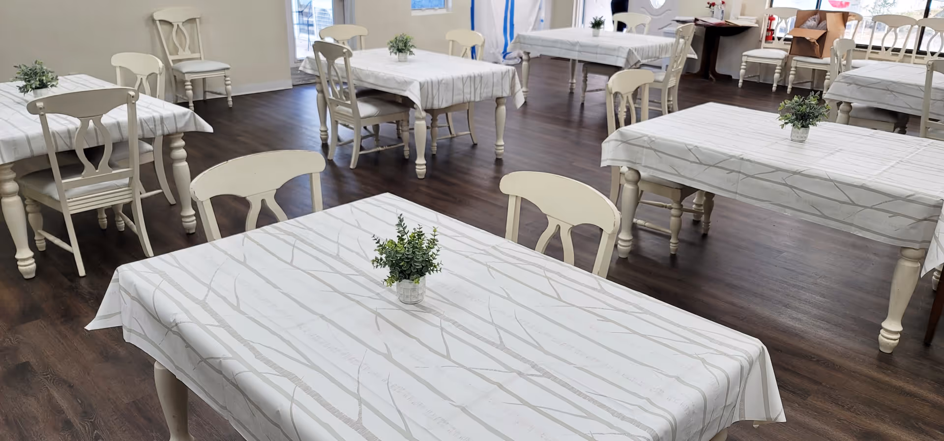 A bright dining room with several rectangular tables covered with white tablecloths featuring a subtle branch pattern. Each table has a small green plant centerpiece. The room has wooden flooring and white chairs with decorative backs. Large windows allow natural light to fill the space.