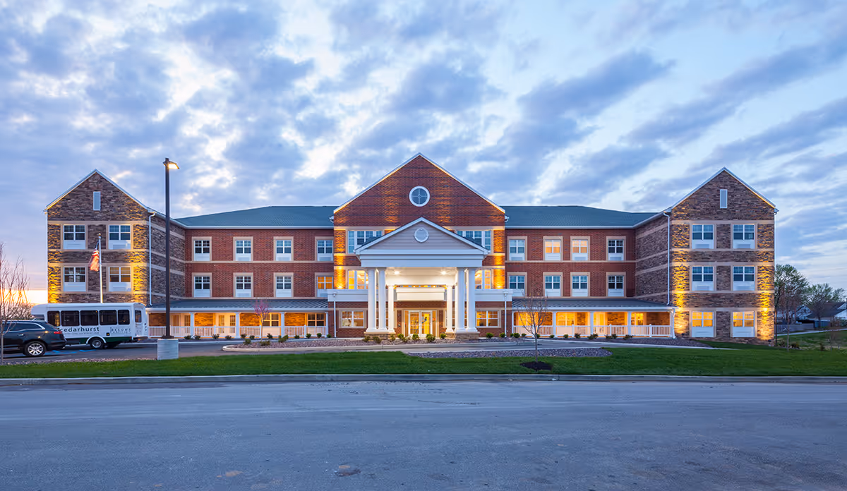 Front exterior view of Cedarhurst Senior Living of St. Charles, a large three-story brick building with white columns at the entrance, illuminated by exterior lights during dusk with a partly cloudy sky.