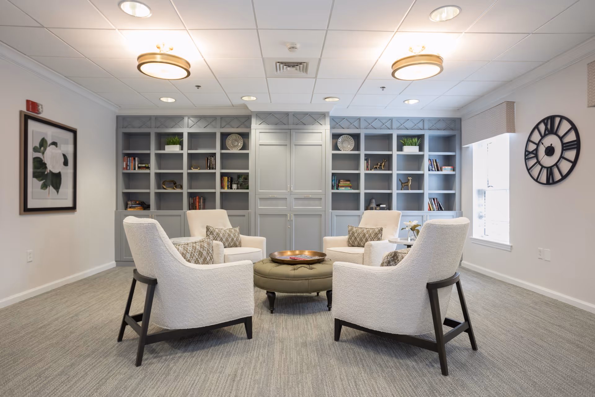 A cozy sitting area in a senior living facility with four white upholstered armchairs arranged around a round green ottoman. Behind the chairs is a large built-in gray bookshelf with decorative items and books. The room has light-colored walls, a large window with blinds, a framed flower artwork on one wall, and a large black clock on another wall. The ceiling has recessed lighting and two round ceiling lights.