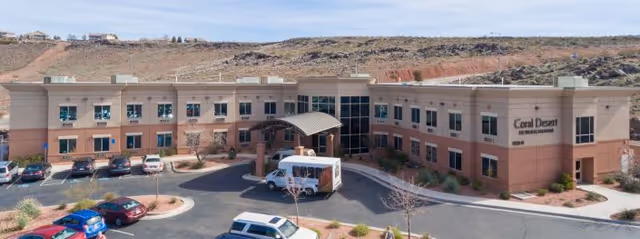 Two-story Coral Desert Rehabilitation And Care building with a covered entrance, parked vehicles in the lot, and desert hills in the background.