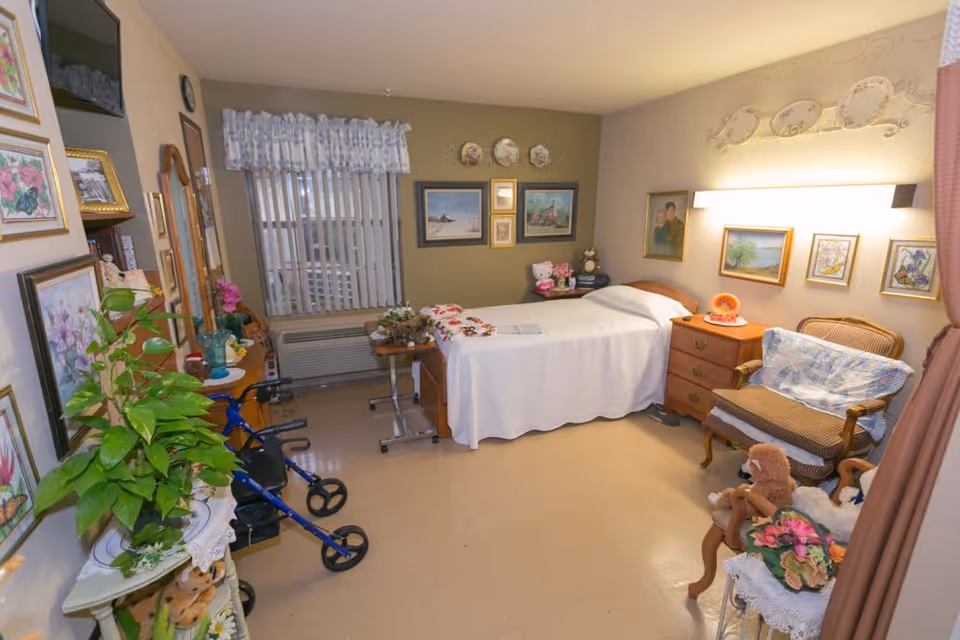 A cozy, well-decorated bedroom in a senior living facility featuring a single bed with a white bedspread, a wooden nightstand with a lamp, a cushioned armchair with a blanket, and various framed pictures and decorative plates on the walls. There is a window with vertical blinds and a floral valance, a blue walker near a small table with plants and flowers, and a small table with stuffed animals and floral decorations.
