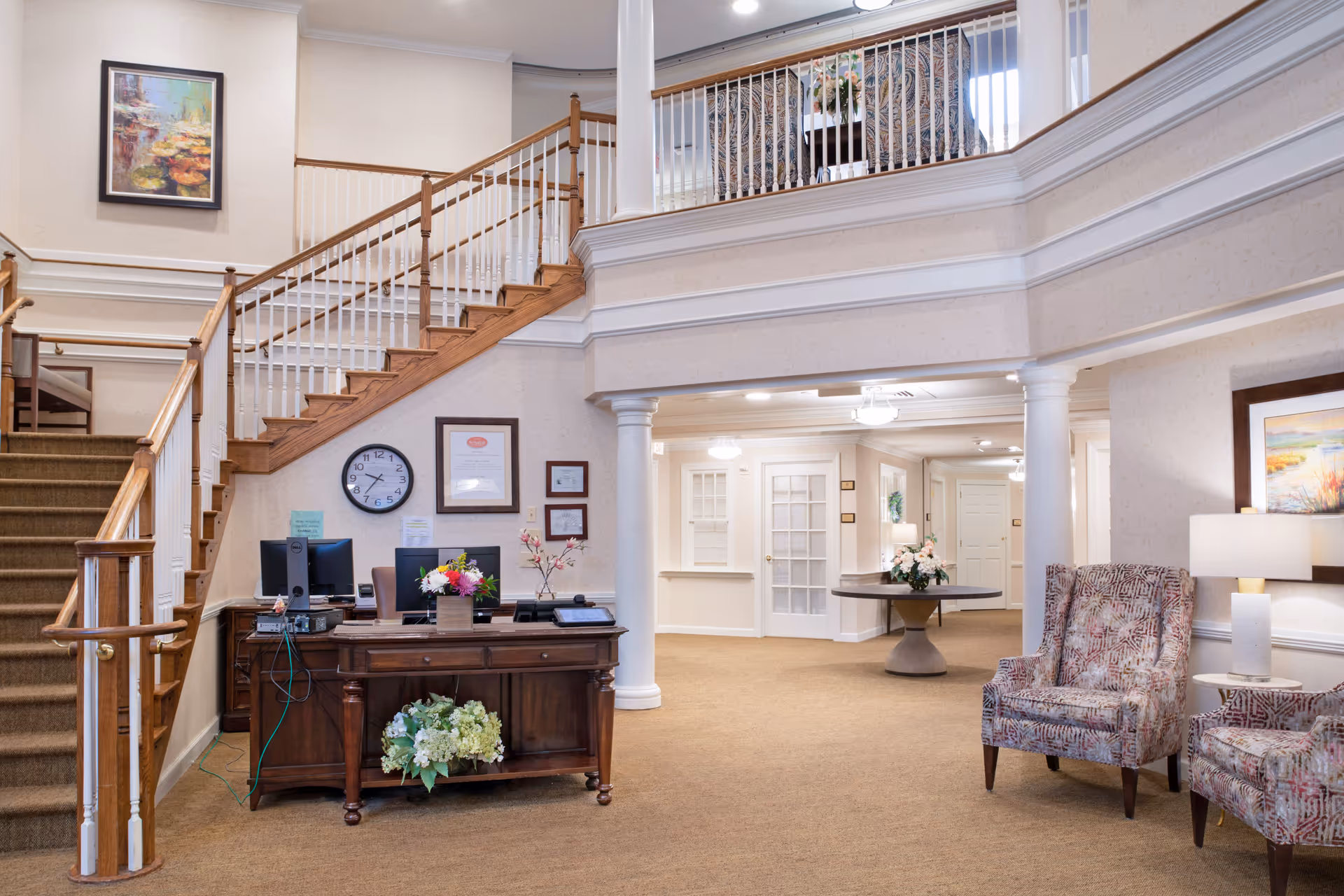 Interior view of a senior living facility lobby with a wooden staircase, a reception desk with computer monitors and flowers, patterned armchairs, a round table with a flower arrangement, and framed artwork on the walls.