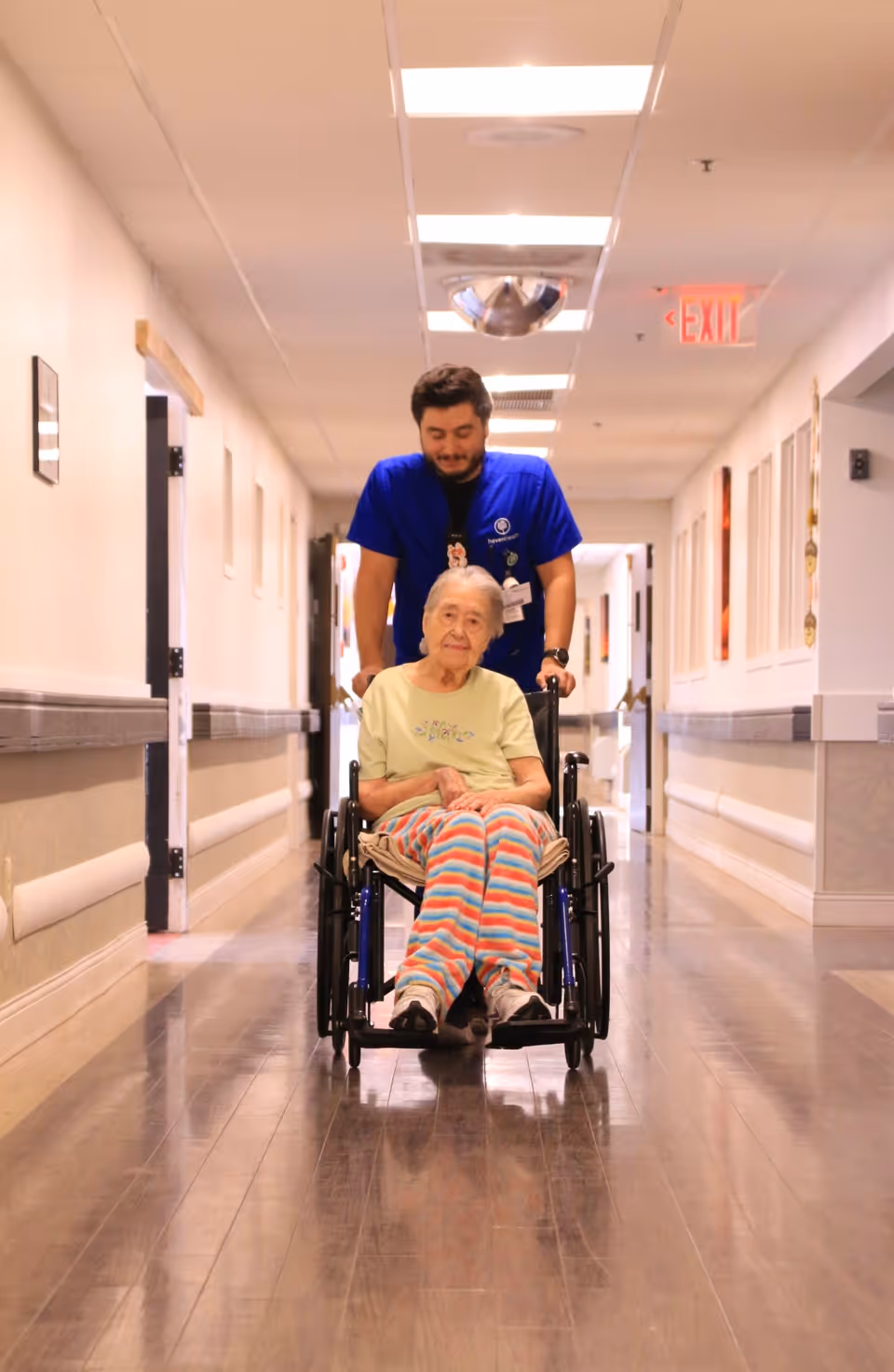 A young male caregiver in a blue uniform is pushing an elderly woman in a wheelchair down a well-lit hallway inside a senior living facility. The elderly woman is wearing a light green shirt and colorful striped pants. The hallway has wooden floors, white walls, handrails, and several doors along the sides.
