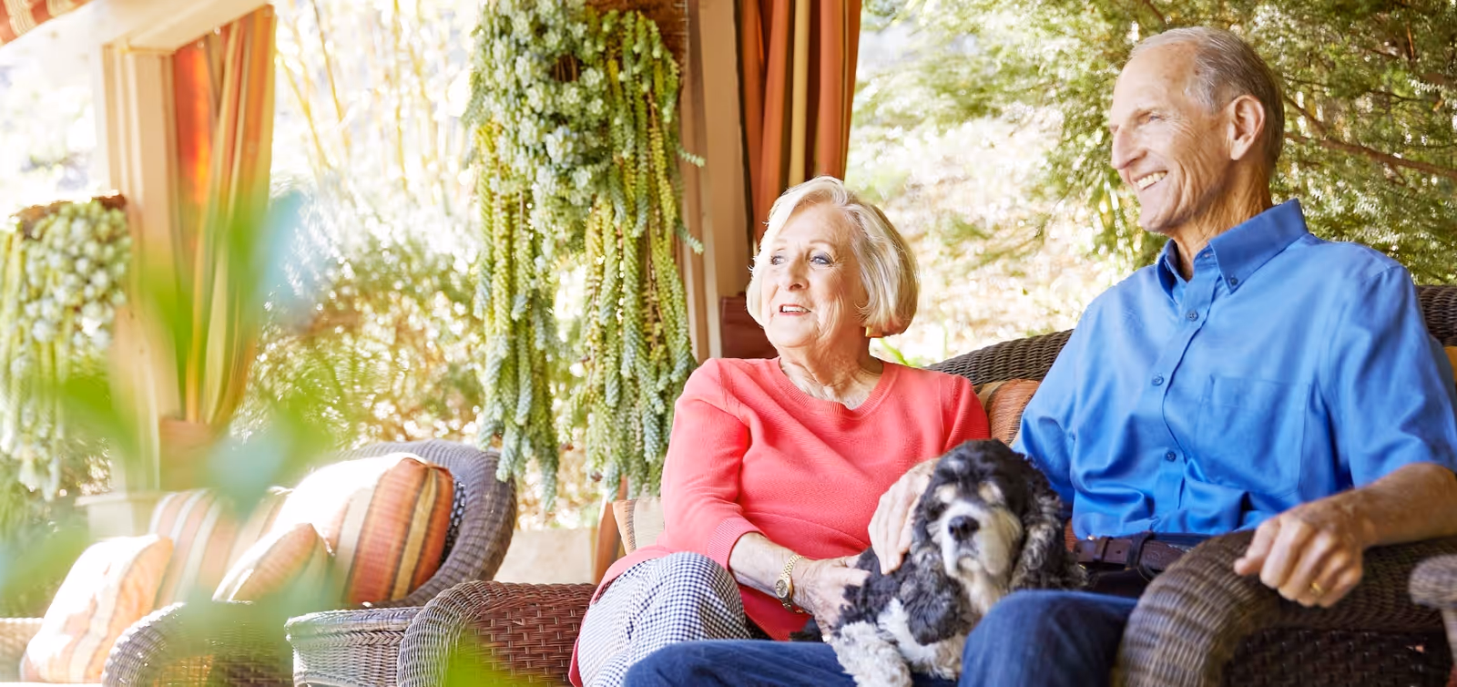 An elderly couple sitting on a wicker sofa on a covered patio, with the woman wearing a red top and the man wearing a blue shirt. They are smiling and holding a black and white dog. There are hanging plants and cushions on the sofa, with greenery visible in the background.