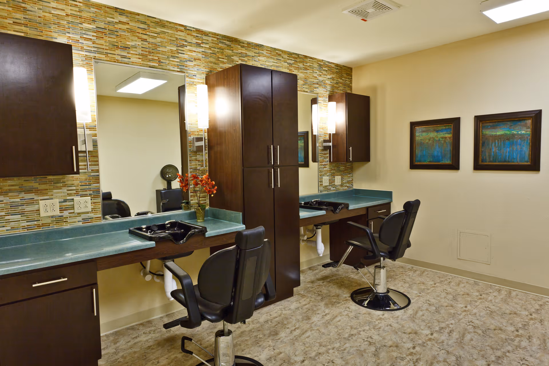 Interior view of a salon area with two black salon chairs in front of two sinks and mirrors. The walls have a decorative tile backsplash behind the sinks and dark wood cabinets above and between the sinks. There are two framed abstract paintings on the adjacent wall and a vase with orange flowers on the counter.