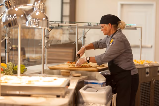 A chef wearing a black hat, glasses, and gloves is slicing food on a large wooden cutting board behind a glass food service counter in a kitchen or cafeteria setting.
