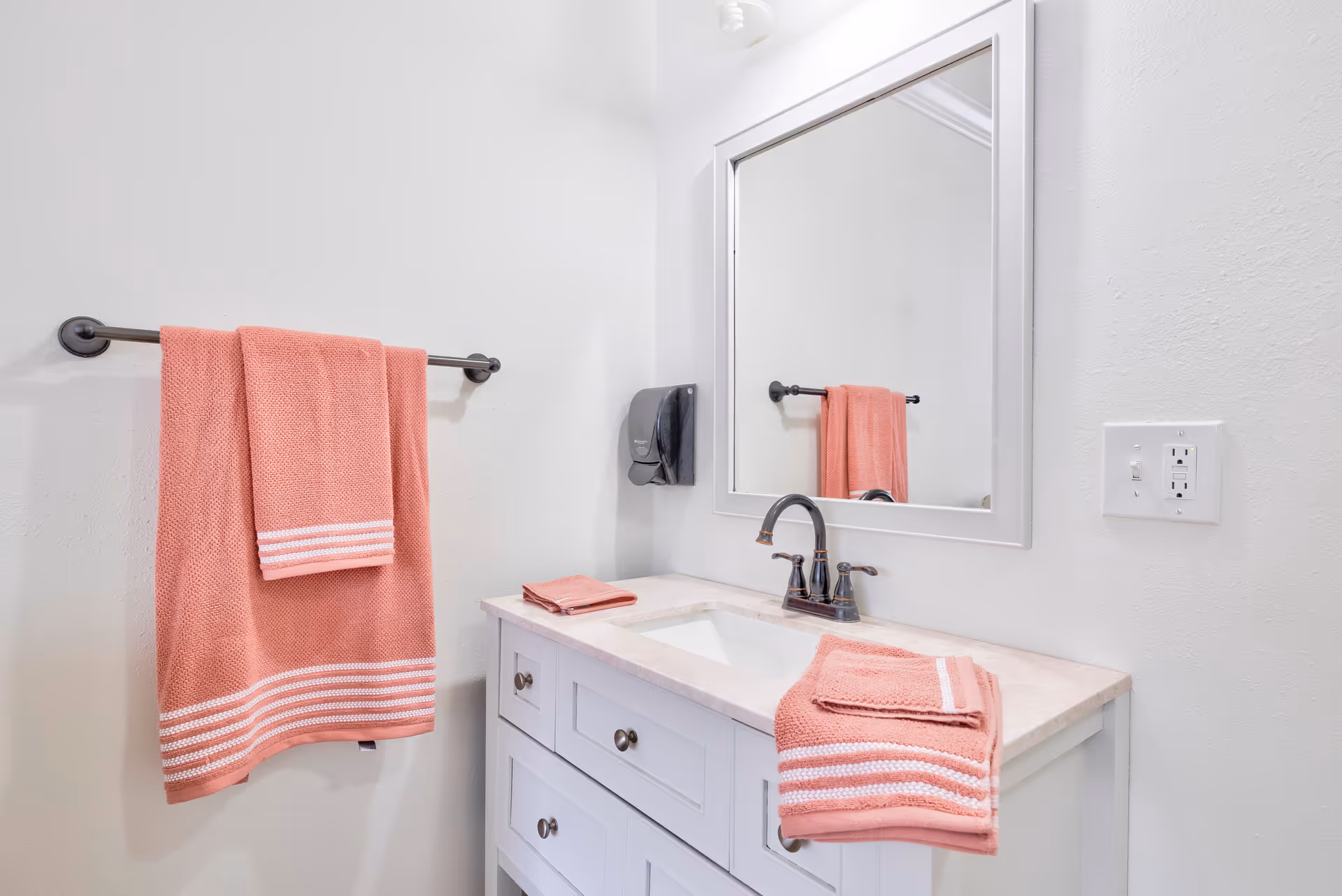A clean bathroom vanity with a white cabinet and beige countertop. There is a rectangular mirror above the sink and a black faucet. Coral-colored towels with white stripes are hanging on a towel rack and placed on the countertop. The walls are painted white.