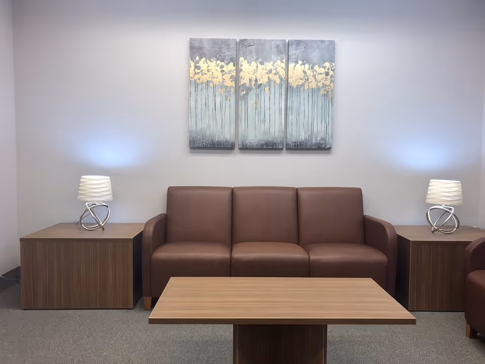 A modern living room with a brown leather sofa centered between two wooden side tables, each holding a decorative lamp with a white shade. Above the sofa, there is a three-panel abstract painting featuring gold and gray tones. In front of the sofa, there is a wooden coffee table. The walls are light gray and the floor is carpeted.