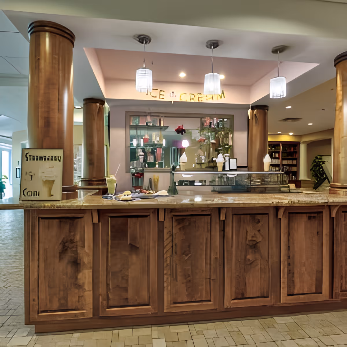 Interior view of an ice cream counter with wooden cabinetry and a granite countertop, featuring a sign that reads 'ICE CREAM' above the counter. There are three pendant lights hanging from the ceiling, and various items including ice cream cones and a menu are displayed on the counter. The background shows a mirrored wall and a glimpse of a lounge area with bookshelves and plants.