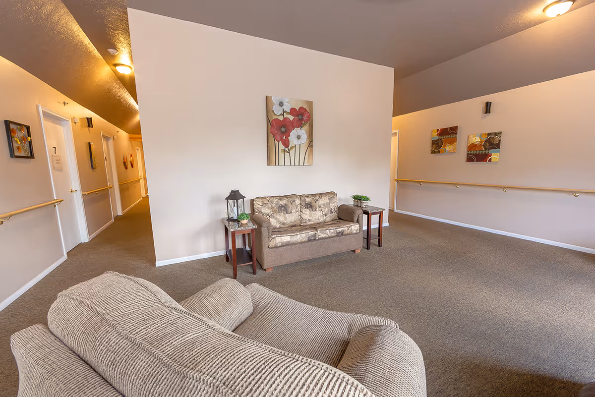 A seating area in a senior living facility hallway with a beige armchair in the foreground and a brown patterned loveseat against a white wall. Two small side tables with decorative plants and a lantern flank the loveseat. The walls are adorned with framed artwork, and handrails run along the hallway walls. The carpet is brown, and ceiling lights provide warm illumination.