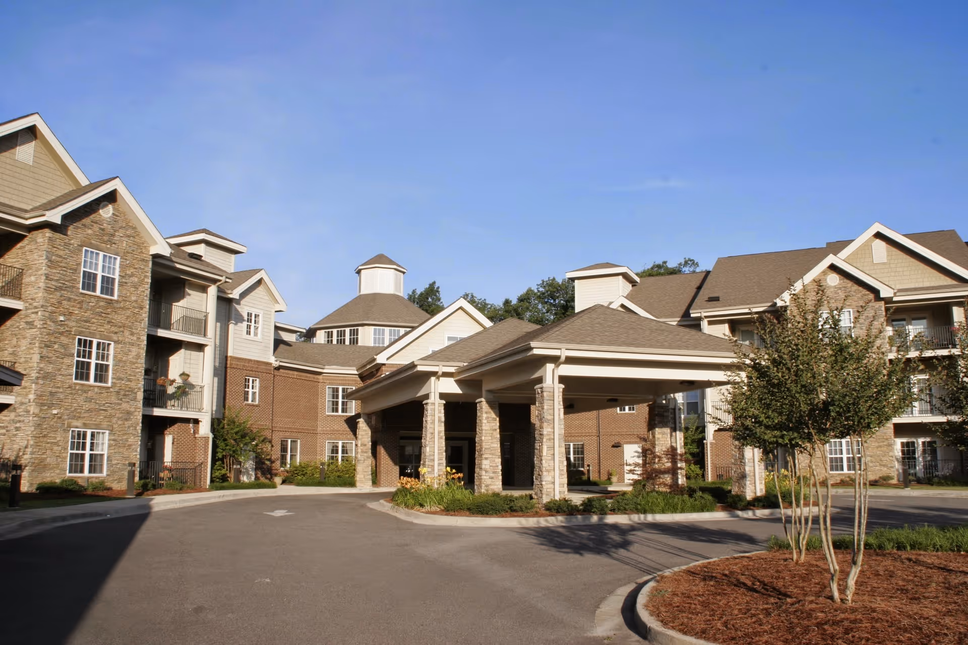 Covered entrance (porte-cochère) and driveway in front of a multi-story senior living building with brick and stone facades under a clear blue sky.