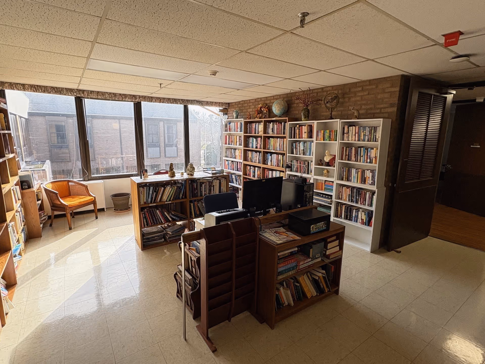 A well-lit room with large windows letting in natural light, filled with multiple bookshelves stocked with books. There is a wooden chair with an orange cushion near the window, a desk with a computer monitor, and various decorative items including globes and small statues on top of the bookshelves. The floor is tiled, and the ceiling has a grid pattern with fluorescent lighting.