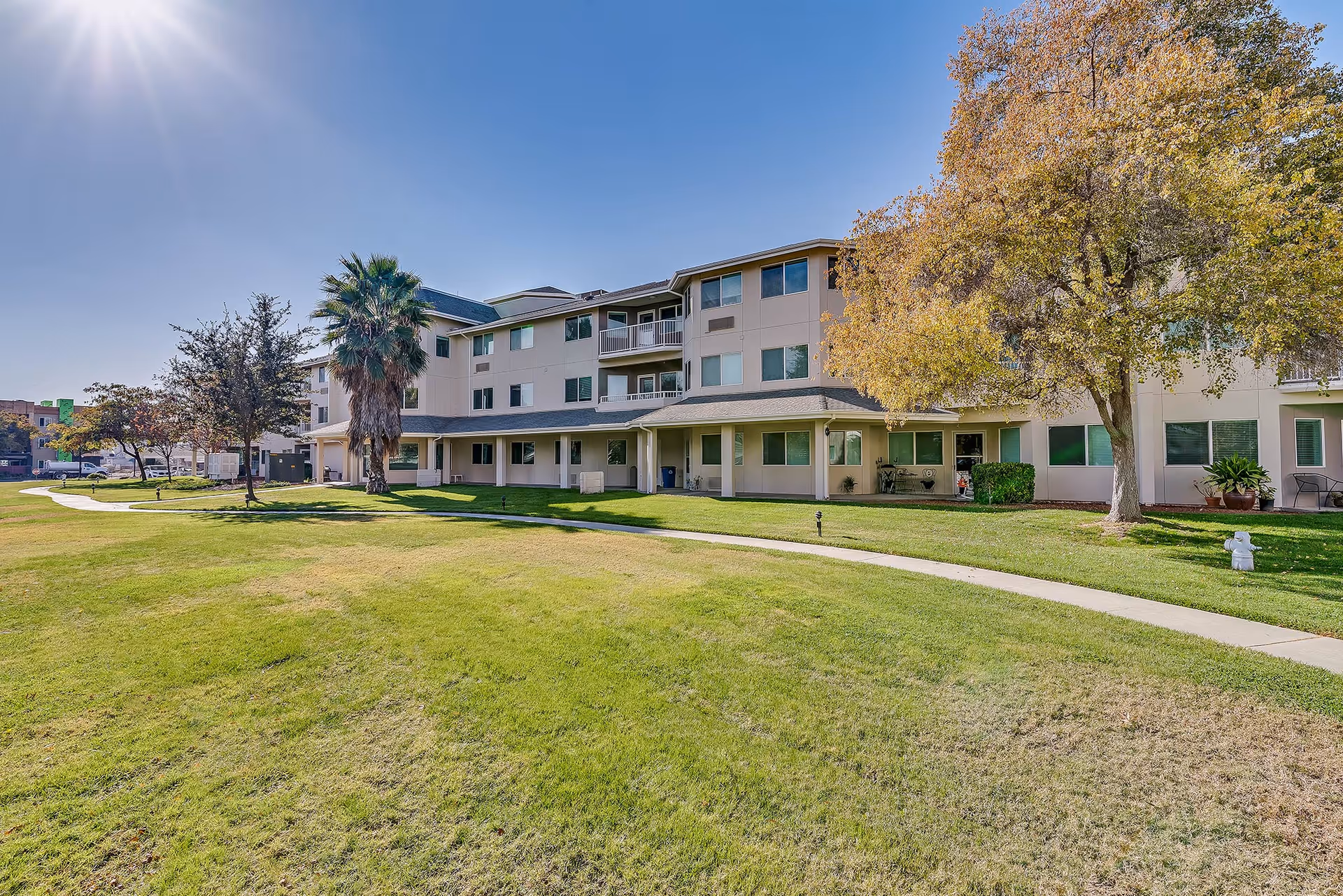 Exterior view of a three-story senior living facility building with a well-maintained green lawn and a paved walkway in front. Trees with autumn foliage and a clear blue sky with the sun shining brightly are visible.