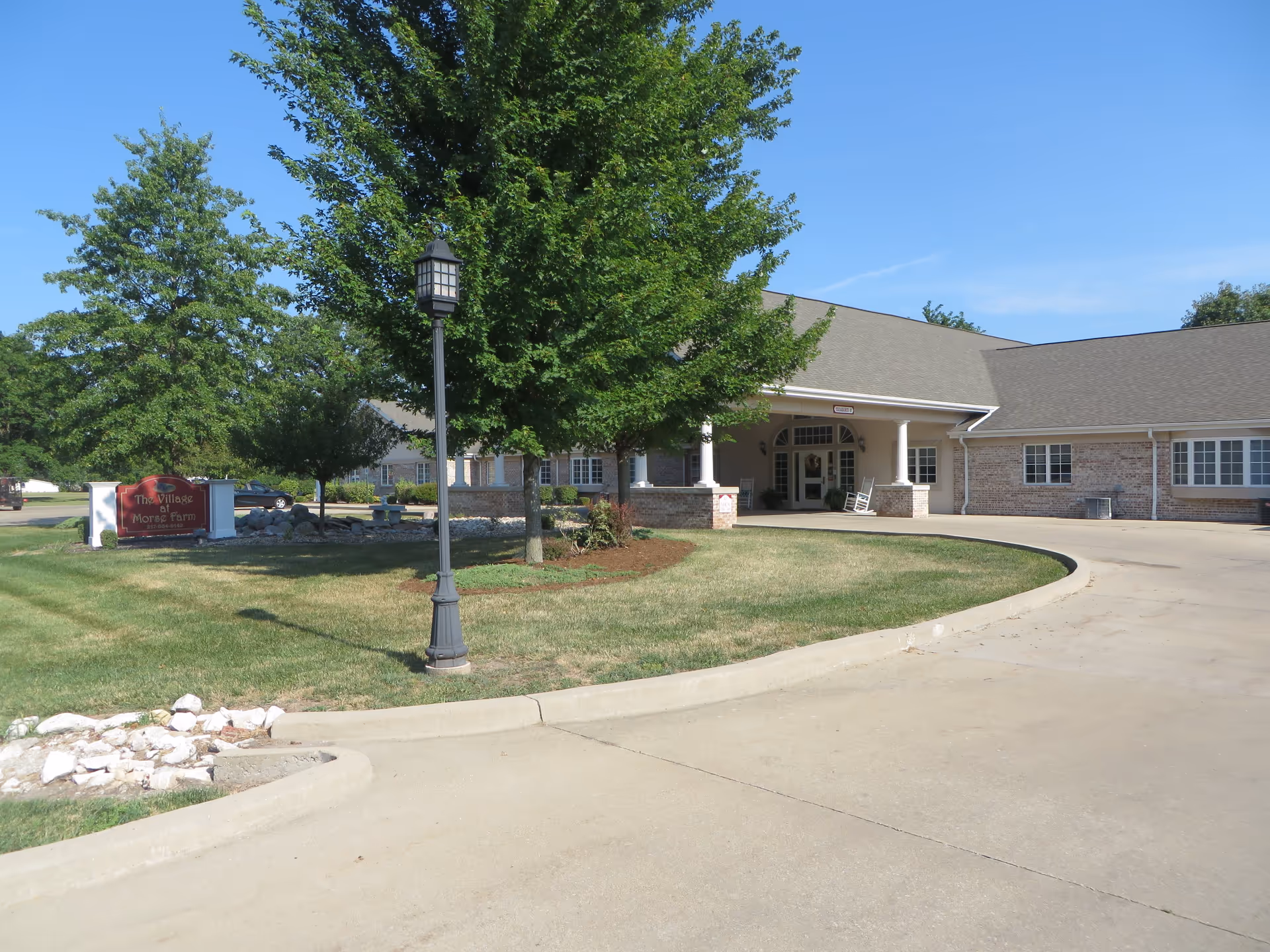 Exterior view of The Village At Morse Farm building with a curved driveway, green lawn, trees, and a lamp post under a clear blue sky.