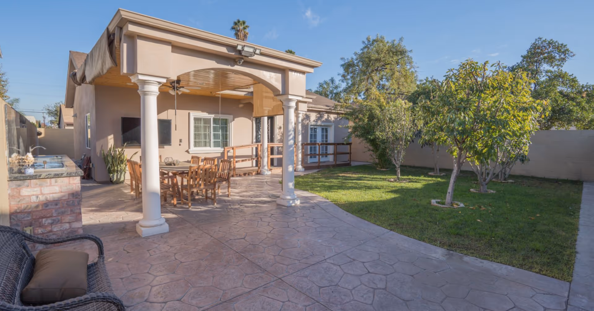 Outdoor patio area of a senior living facility with a covered seating area featuring wooden chairs and a table, a mounted TV on the wall, a small outdoor kitchen with a sink and countertop, and a grassy yard with several small trees enclosed by a beige wall.