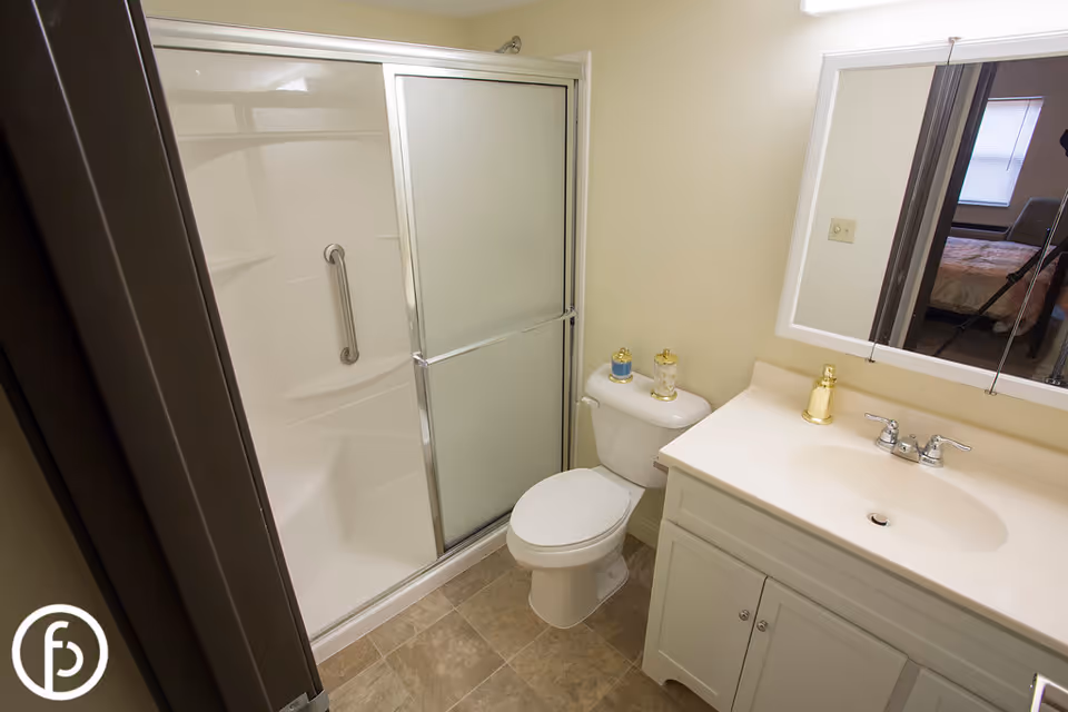 A clean bathroom featuring a white toilet, a vanity with a sink and gold-colored faucet, a large mirror above the sink, and a shower with sliding frosted glass doors and a grab bar inside. The floor is tiled, and a bedroom is partially visible through the mirror reflection.
