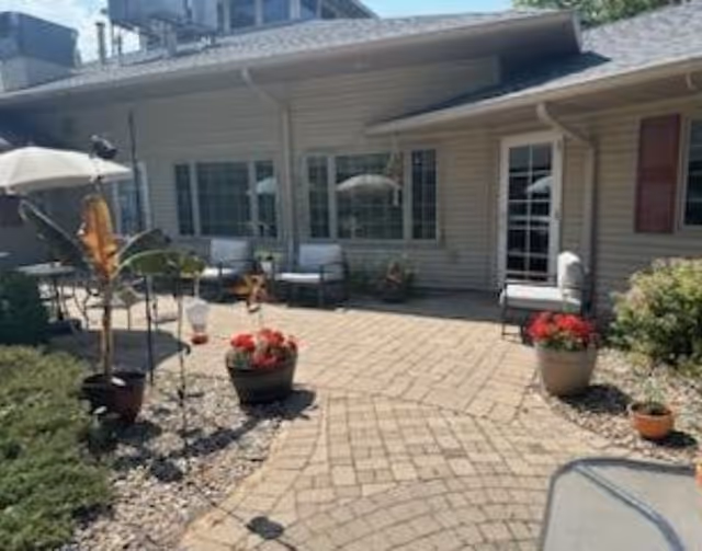 Paved outdoor patio courtyard with potted flowers, seating, umbrellas, and the building's windows and door.
