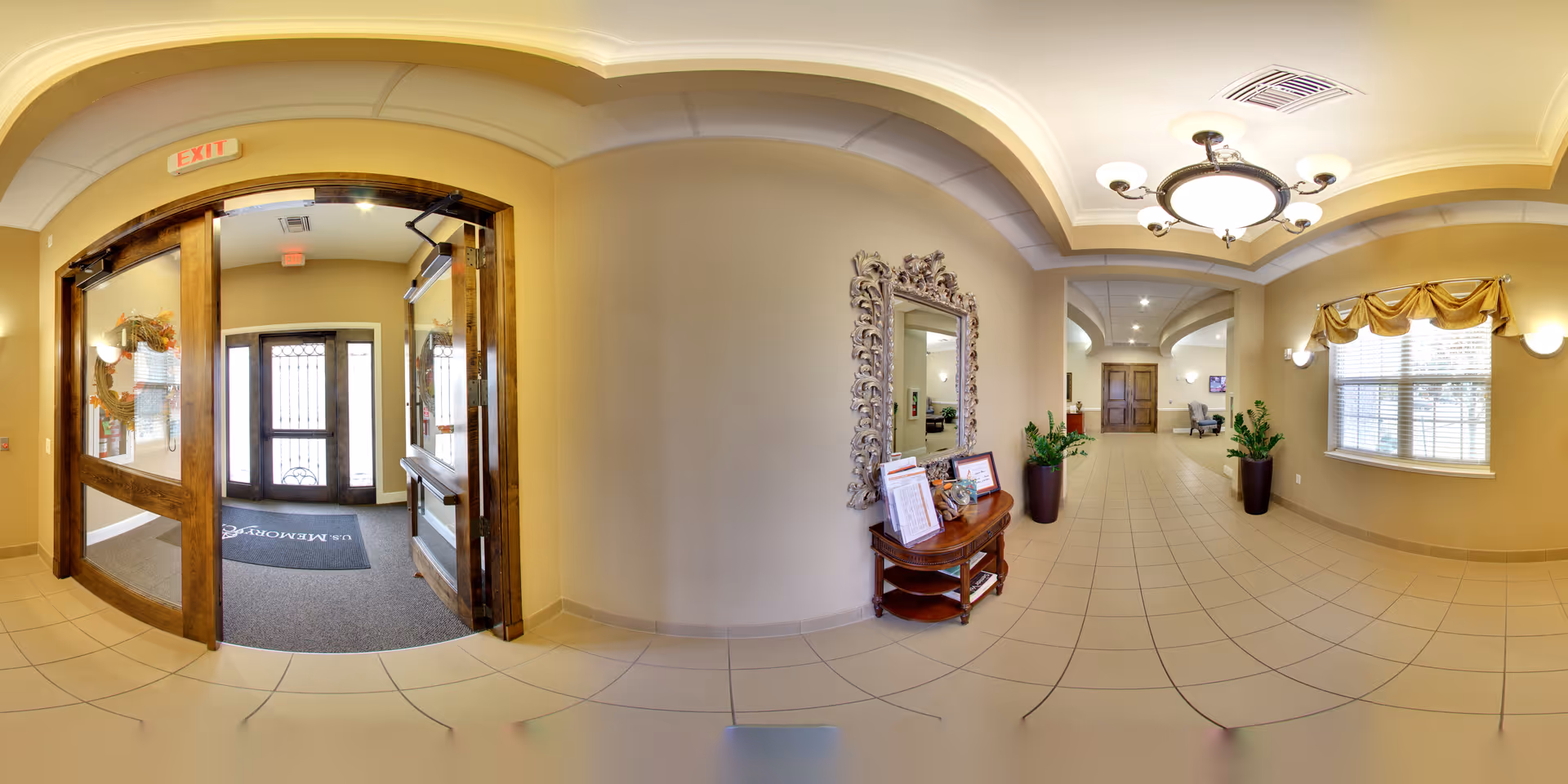 A spacious and well-lit hallway in a senior living facility with beige tiled floors and cream-colored walls. The hallway features a large decorative mirror above a wooden table holding brochures and a small plant. There are two large potted plants along the wall, a window with a yellow valance, and a ceiling light fixture. Double wooden doors with glass panels lead to an entrance area with a welcome mat and an exit sign above the doors.