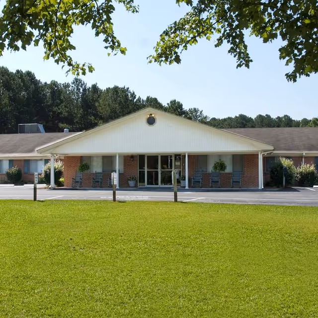 Front exterior view of a single-story brick building with a white gabled roof and a covered entrance. There are several rocking chairs and potted plants on the porch. The building is surrounded by a well-maintained green lawn and trees in the background under a clear blue sky.