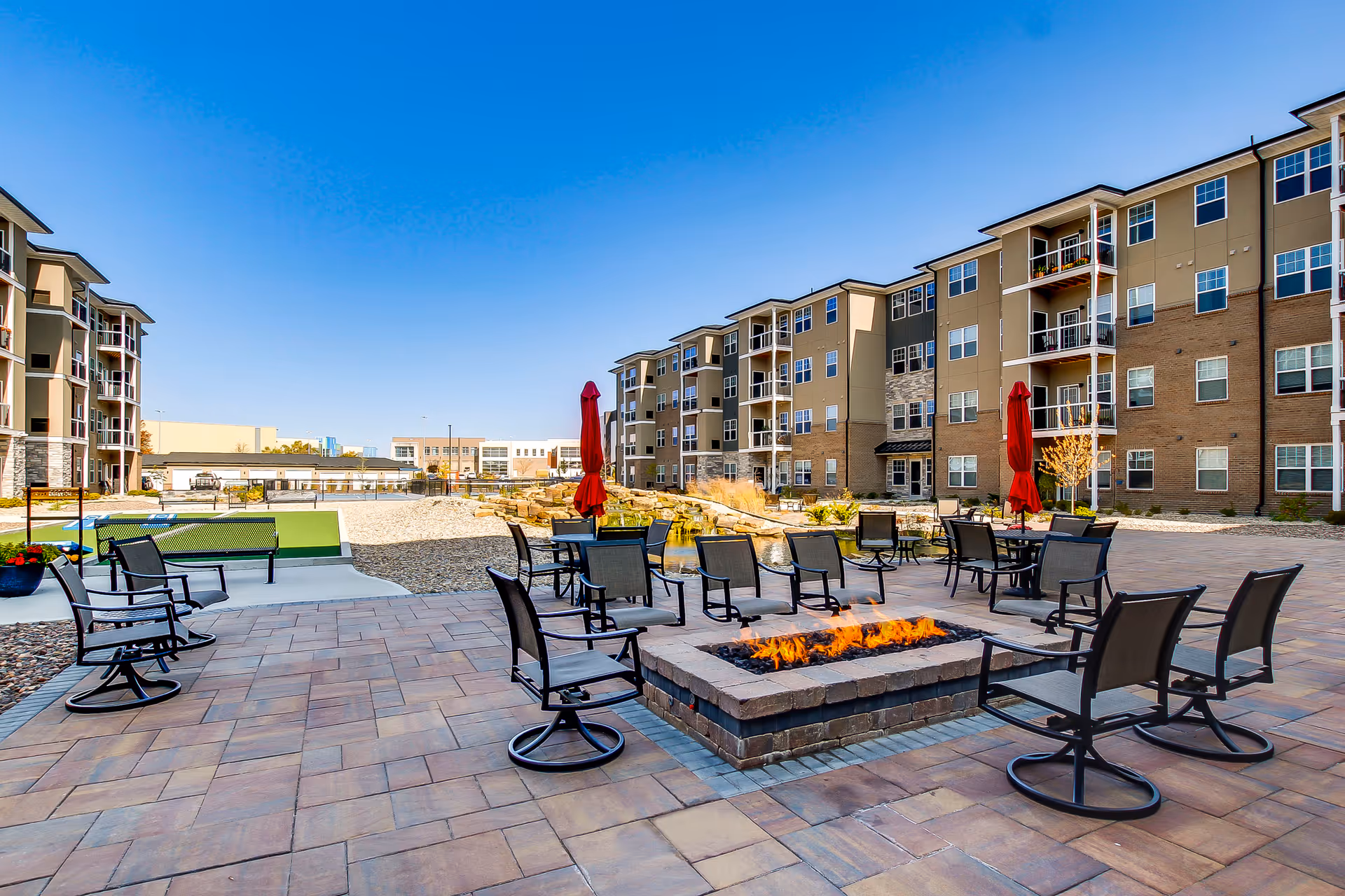 Outdoor patio area at Encore 55+ Boutique Apartments in Plainfield featuring a rectangular fire pit surrounded by multiple black metal chairs with mesh seats and backs. The patio is paved with large stone tiles and includes several tables with closed red umbrellas. In the background, there are multi-story apartment buildings with balconies under a clear blue sky.