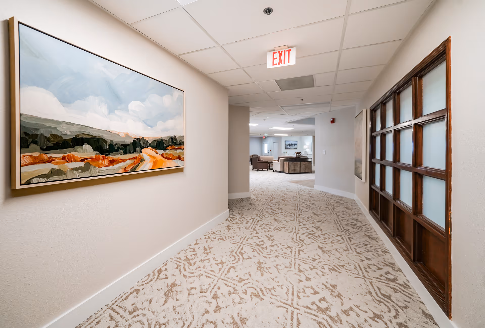 A carpeted hallway in a senior living facility with a large landscape painting on the left wall and a wooden-framed glass window on the right. The hallway leads to a common area with seating and another piece of artwork on the far wall. An exit sign is visible on the ceiling.
