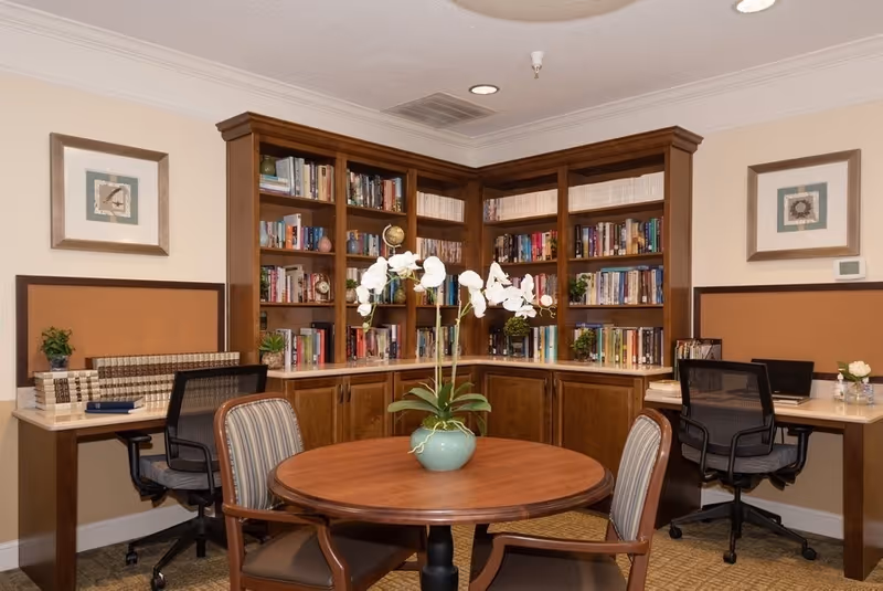 A cozy reading and work area in an assisted living facility featuring a round wooden table with a potted white orchid in the center, surrounded by four chairs. Behind the table is a built-in wooden bookshelf filled with books and decorative items. On either side of the bookshelf are two workstations with office chairs, books, and small plants. The walls have framed artwork and a thermostat.