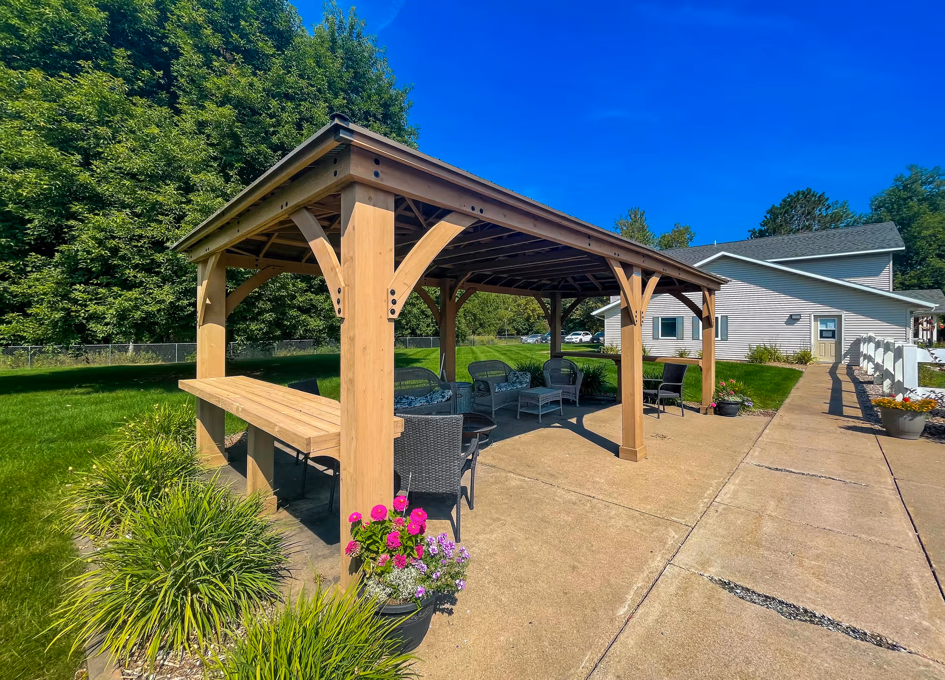 Outdoor wooden gazebo with seating area including chairs and a table, surrounded by green grass and plants with pink flowers. A paved walkway leads to a light-colored building in the background under a clear blue sky.