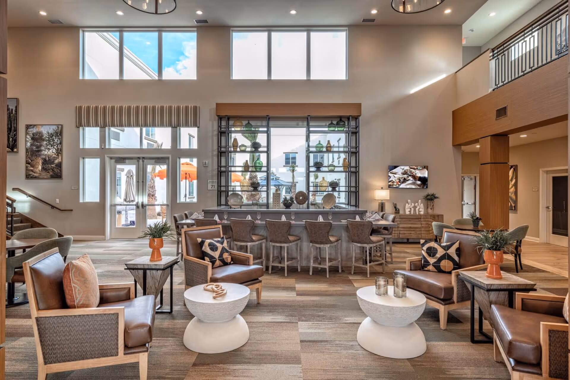 A spacious and well-lit senior living facility common area with high ceilings and large windows. The room features comfortable leather armchairs with decorative pillows, small round tables with plants and candles, and a long dining table with chairs. There is a decorative shelving unit with vases and plants, a flat-screen TV mounted on the wall, and modern lighting fixtures. The overall decor is warm and inviting with neutral tones and wood accents.