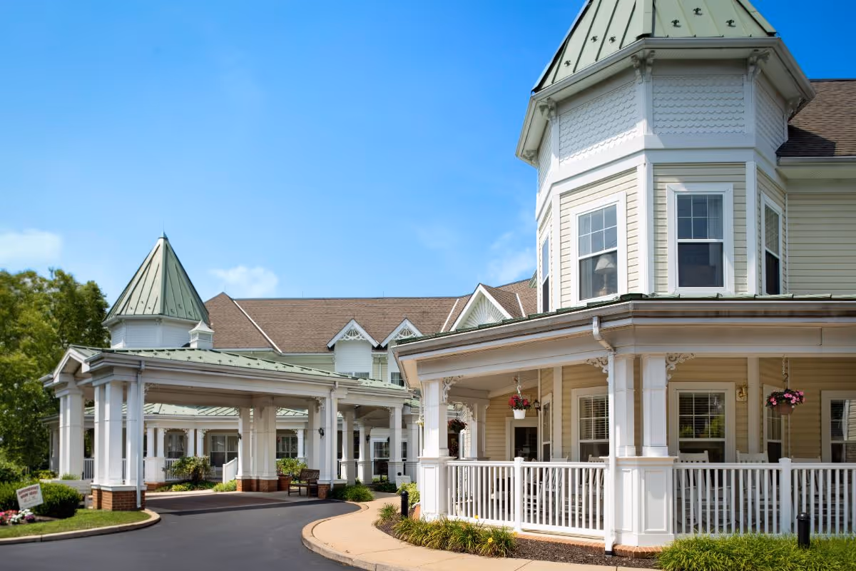 Exterior view of a senior living facility building with a covered entrance, white columns, and a wrap-around porch with hanging flower pots. The building has a light yellow exterior with white trim and a green metal roof. The sky is clear and blue.