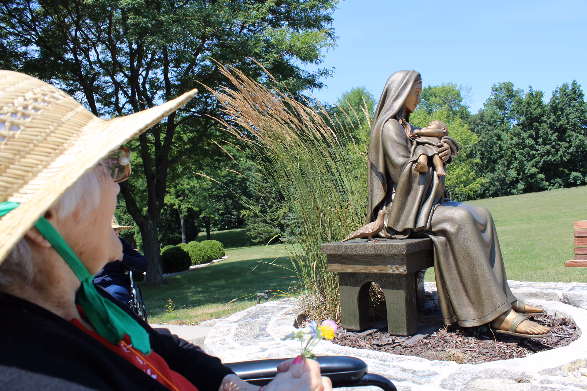 An elderly woman in a straw hat looks toward a bronze statue of a seated mother holding a child in a grassy outdoor garden.