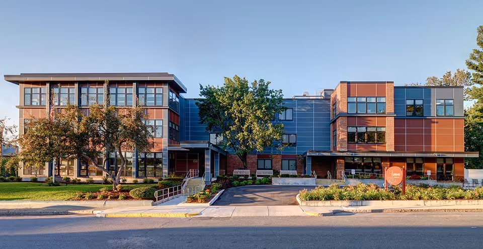 Front exterior view of a modern three-story building with large windows, trees, and landscaped greenery in front under a clear blue sky.