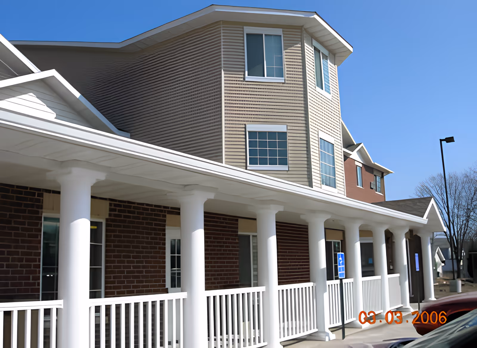 Exterior view of a multi-story building with beige and brown siding, white columns supporting a covered porch, and several windows. There is a clear blue sky above and a parking area with cars in front. A handicap parking sign is visible near the porch.
