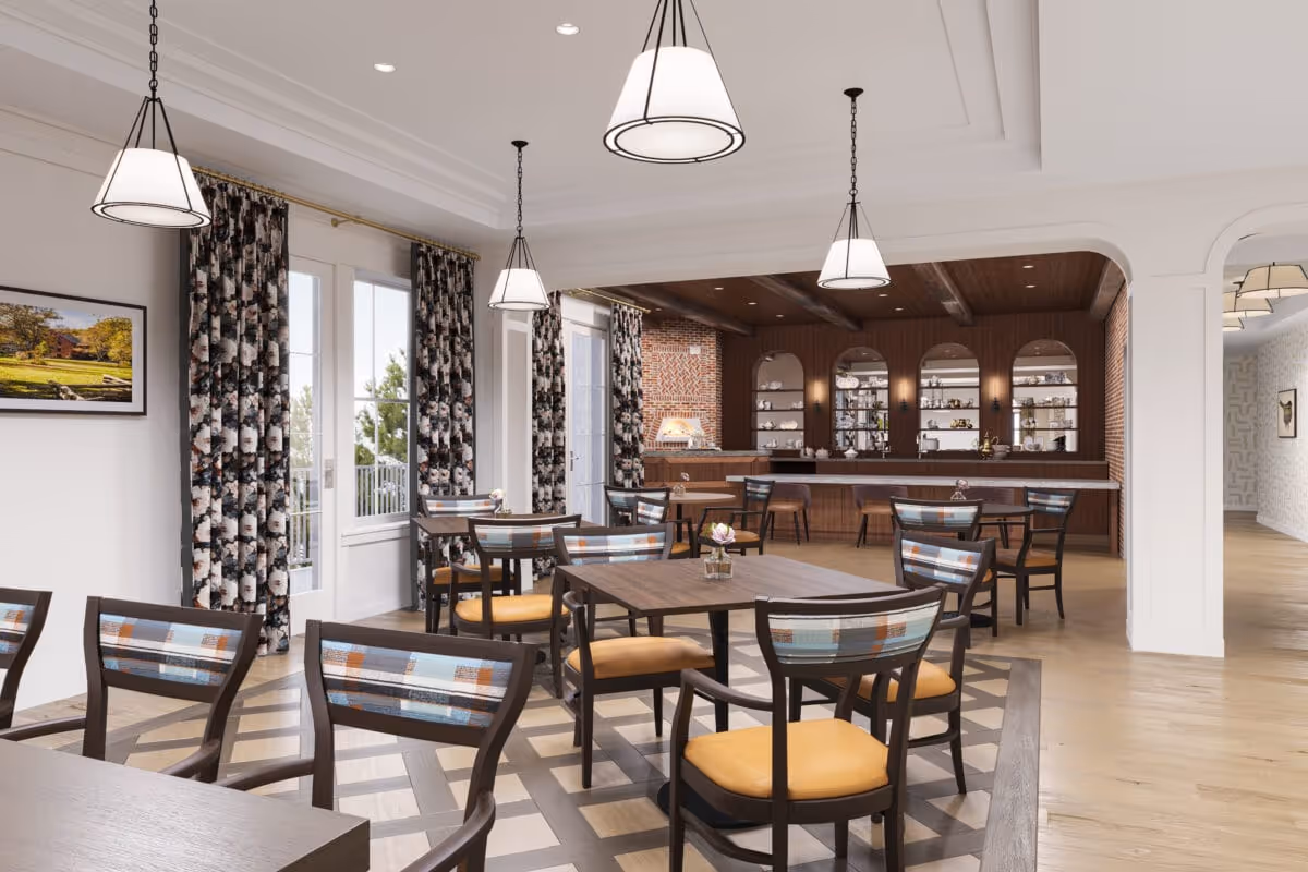 A bright dining room with several wooden tables and chairs featuring patterned backs and yellow cushions. The room has large windows with floral curtains, pendant lights hanging from the ceiling, and a bar area with shelves and a brick oven in the background.