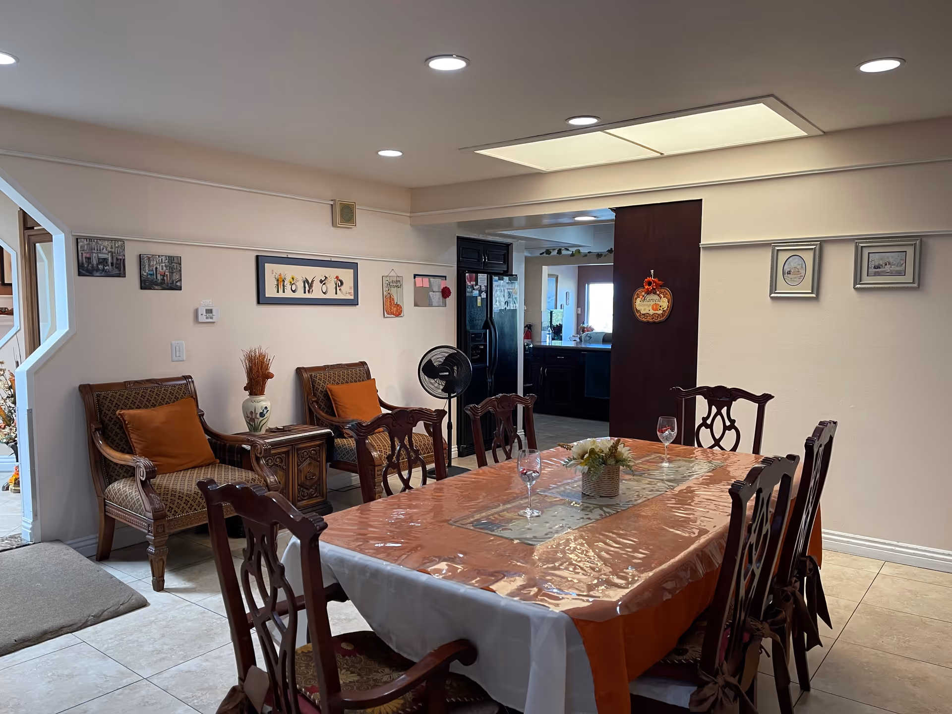 A dining room with a long table covered with a plastic tablecloth and an orange runner. The table is set with three wine glasses and a small flower arrangement centerpiece. There are eight wooden chairs around the table. In the background, there are two armchairs with orange cushions and a small side table with a vase. The walls are decorated with framed pictures and a thermostat. The room is well-lit with recessed ceiling lights and a large ceiling light panel. A kitchen area is visible through an open doorway.