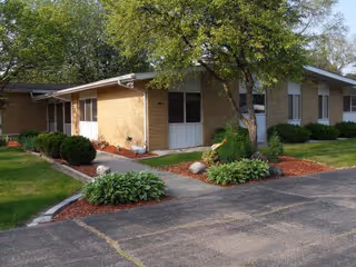 Single-story brick building with a sloped roof surrounded by green grass, bushes, and trees. A paved driveway and walkway lead to the entrance, with landscaping including mulch and plants along the path.