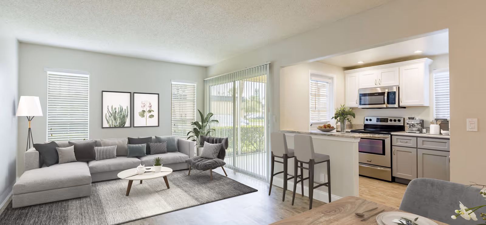 A bright and modern living room and kitchen area in a senior living facility. The living room features a large gray sectional sofa with multiple cushions, a small oval coffee table with a teapot and plant, a floor lamp, and two framed botanical prints on the wall. Large windows with blinds and a sliding glass door with vertical blinds allow natural light to fill the space. The kitchen has white upper cabinets, gray lower cabinets, stainless steel appliances including a microwave and stove, and a breakfast bar with two high chairs. A dining table with place settings is partially visible in the foreground.