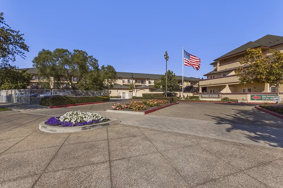 Driveway and landscaped front entrance of a senior living facility with an American flag and multi-story building.