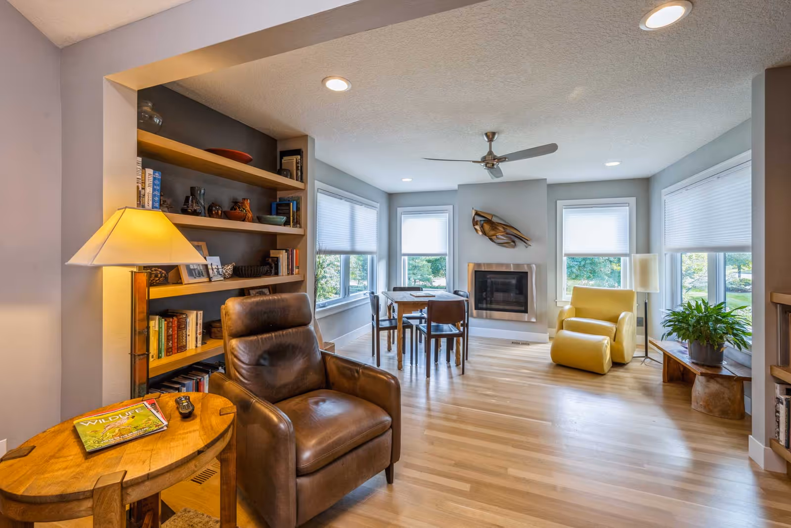 Bright, modern living room with a leather armchair, built-in bookshelves, fireplace, dining table, and a yellow lounge chair by large windows.